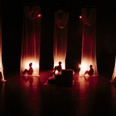 Five dancers are silhouetted behind tall, sheer curtains, each lit by a soft, warm glow.