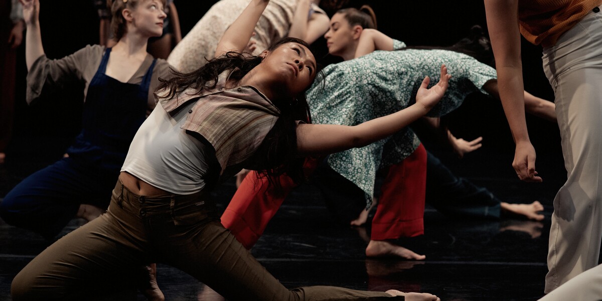 six dancers stretch and look in different directions wearing everyday clothes. One dancer is in the foreground in a deep lunge and their arms stretched to the sky