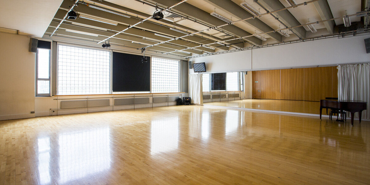 A dance studio picture taken from the corner of the room. It has a wooden floor and a mirror on one of the walls There is also a piano in the right of the picture.