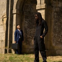 Image of two dancers standing in front of a church. One is in the background dressed in blue, standing against the wall. The other is dressed in black and is slightly more in the foreground. They look pensive and lost in thought.