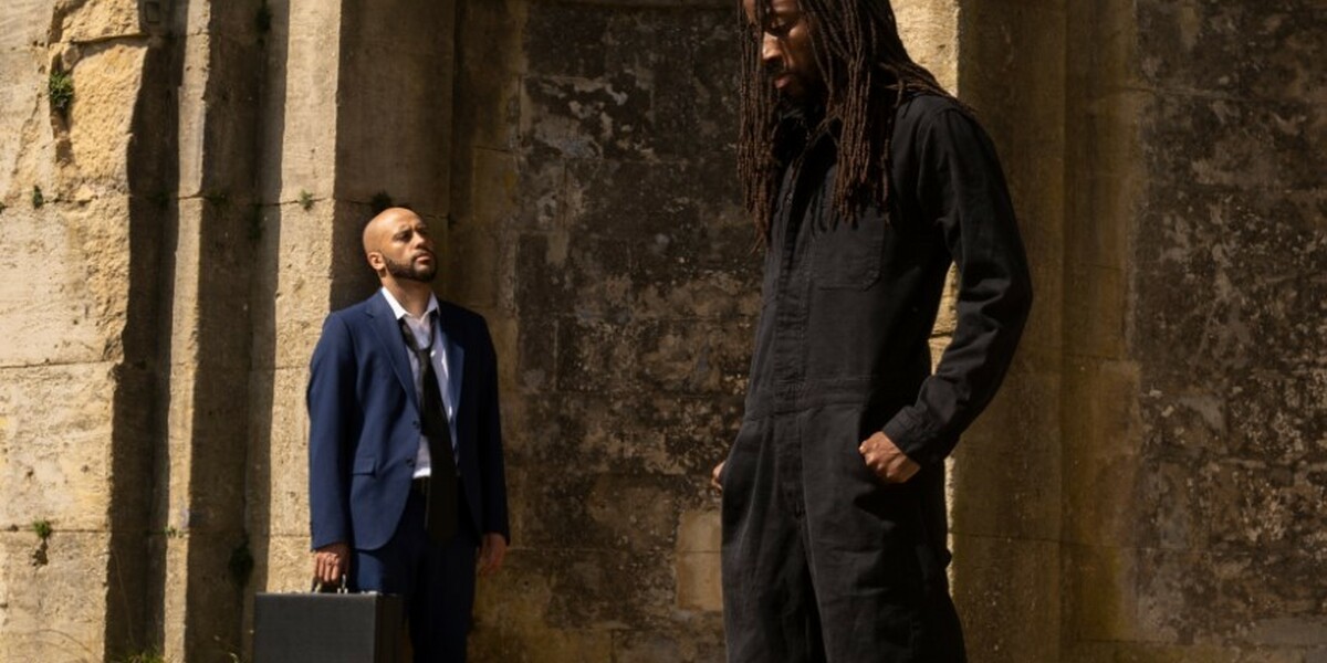 Image of two dancers standing in front of a church. One is in the background dressed in blue, standing against the wall. The other is dressed in black and is slightly more in the foreground. They look pensive and lost in thought.