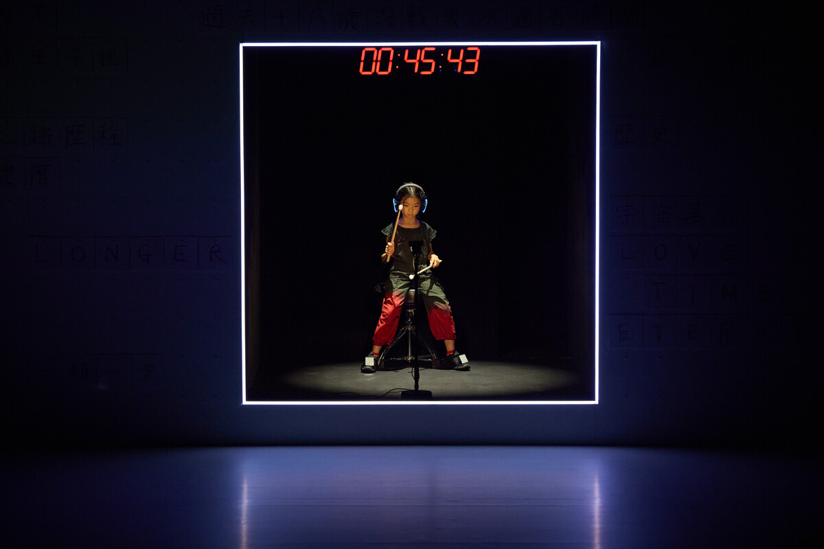 A photo of a child performer sat in a box with headphones on playing a motion sensor drumkit. There is a red digital clock above them.