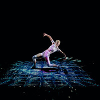 A dancer moves robotically across an illuminated grid floating in space. They wear futuristic white clothing, standing in a powerful stance, their arms outstretched as they lunge forwards.