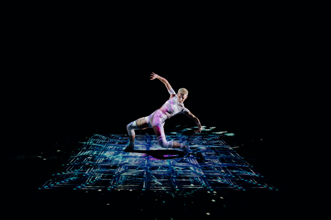 A dancer moves robotically across an illuminated grid floating in space. They wear futuristic white clothing, standing in a powerful stance, their arms outstretched as they lunge forwards.