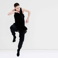 A dancer dressed in black with short dark hair is in the frame. The background is a white cream colour. The dancer jumps into the air, their right leg slightly raised as if they are running in the air. Their right arm is bent inwards towards the chest so the front of the hand touches their chin. The other arm is bent as well but is more to the side.