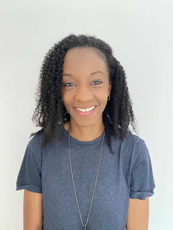 A woman wearing a blue short sleeved t-shirt and a long necklace smiles at the camera, there is a cream background behind her.