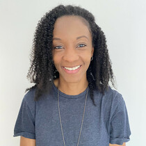 A woman wearing a blue short sleeved t-shirt and a long necklace smiles at the camera, there is a cream background behind her.