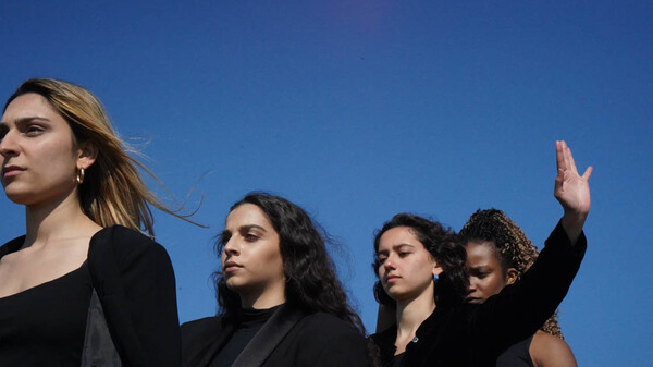 Four women stand in a line on the diagonal, they are looking into the far distance and there is lots of bright blue sky above them