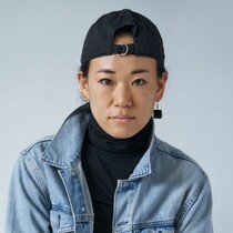 Yukiko is looking in the camera. She is wearing a black cap backwards, a darl grey polar neck t-shirt and a denim jacket.