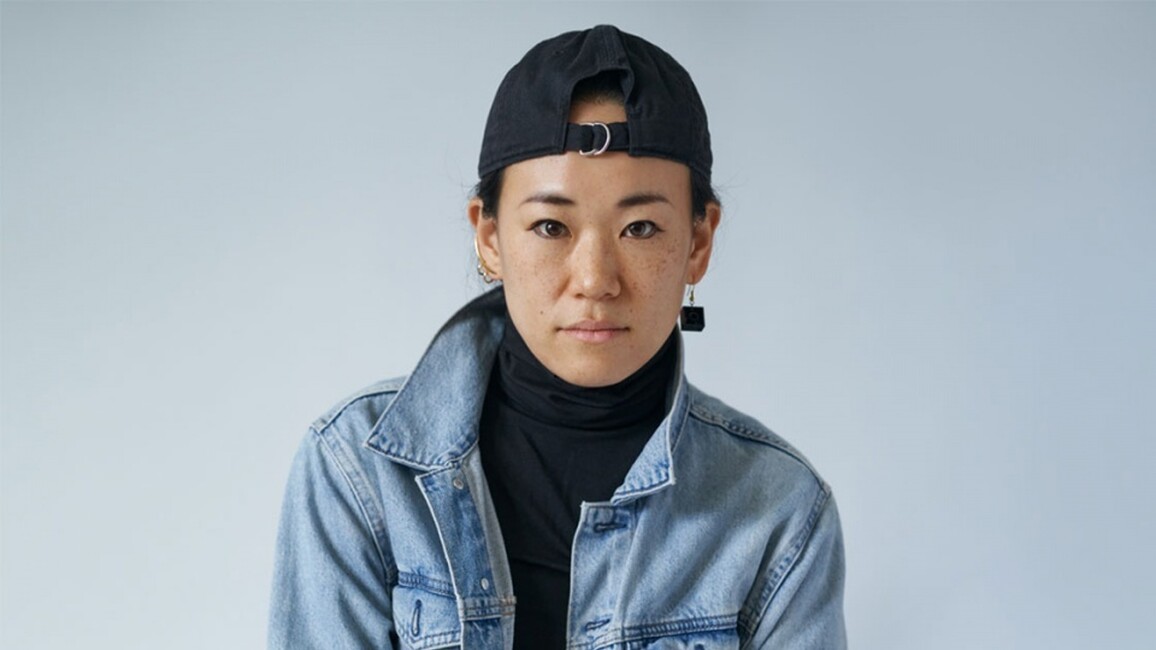 Yukiko is looking in the camera. She is wearing a black cap backwards, a darl grey polar neck t-shirt and a denim jacket.