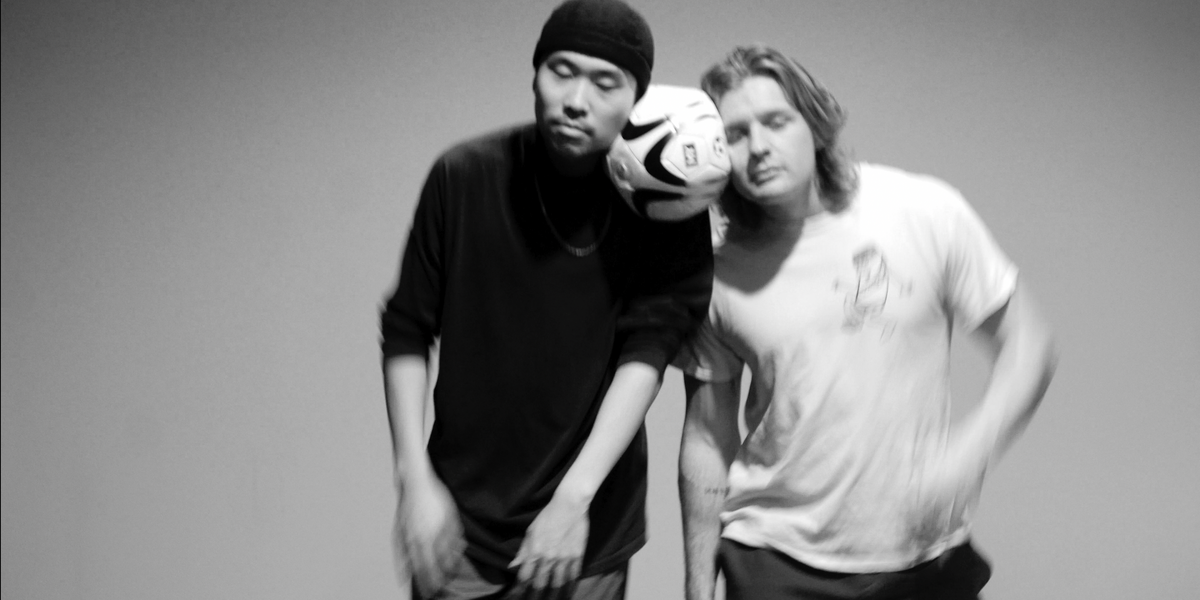 Image of two dancers. They hold a football between their heads. One dancer is wearing black and has a black hat on. The other is wearing a white graphic t-shirt.