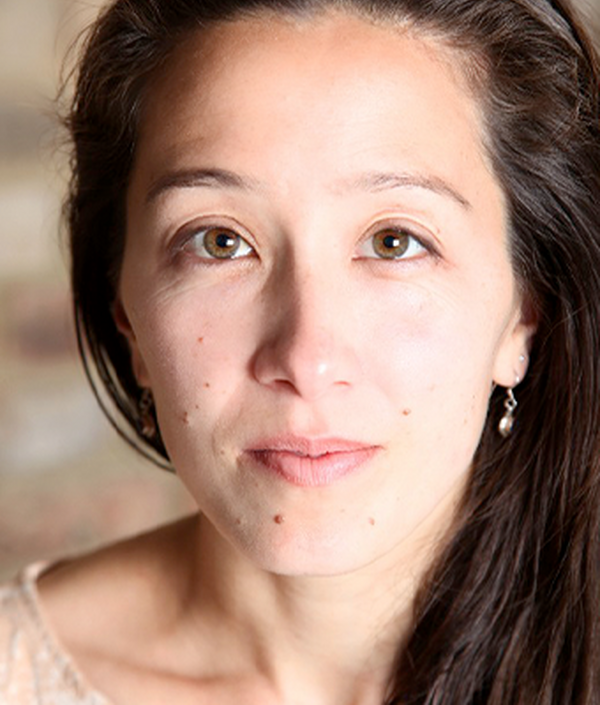 A woman with brown eyes looks directly into the camera, she is wearing a cream top and had brown long hair