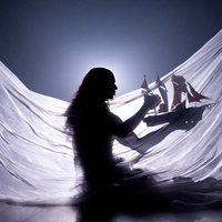 Silhouette of Little M, a person with long hair, wearing a draped flowing translucent fabric, holding a model ship with red sails. Backlit to create a dreamy, underwater-like glow, the image evokes a sense of a dreamlike world.