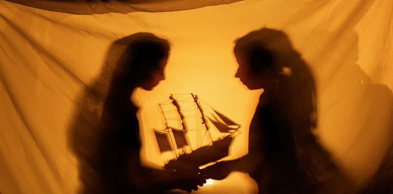 Two silhouetted figures of young people within a warm, golden fort holding a model ship together. Their soft shadows and the ship’s outline create a dreamlike moment that suggests storytelling.