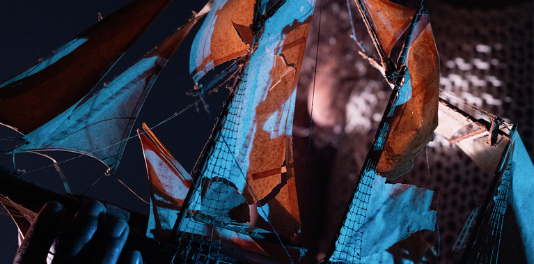 Hands holding a model boat, with sails backlit in blues and a light touch of red highlighting the worn textures of the wood and fabric of the sails