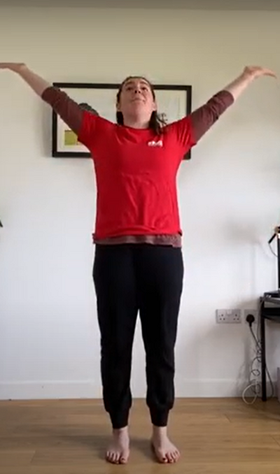 Dance teacher in their lounge wearing a red t-shirt and black trousers stretching their arms upwards towards the ceiling.