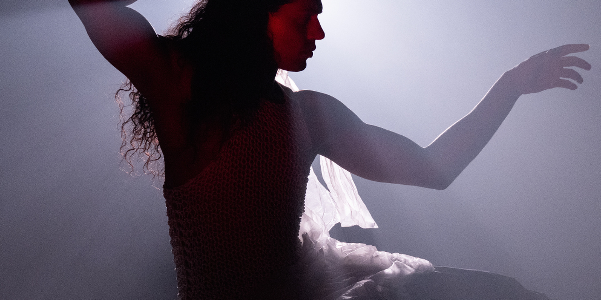 Silhouette of Little M, a person with long hair, posed wearing a flowing, netted costume. They hold a model ship above their head as light beams from behind create an ethereal, oceanic glow. The image evokes themes of transformation, dream like wonder, and the magical world below the sea.