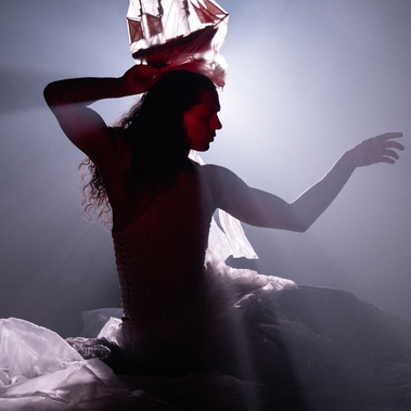 Silhouette of Little M, a person with long hair, posed wearing a flowing, netted costume. They hold a model ship above their head as light beams from behind create an ethereal, oceanic glow. The image evokes themes of transformation, dream like wonder, and the magical world below the sea.