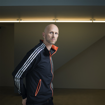 Image of Wayne Mc Gregor. In the background of the image is a thin light beam, which runs across the image. Wayne is in the foreground and he wears a tracksuit.